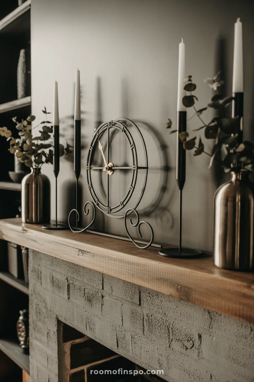 Rustic wood beam mantel over a dark brick fireplace, featuring a wrought iron clock, tall taper candles, and brass vases with eucalyptus sprigs.
