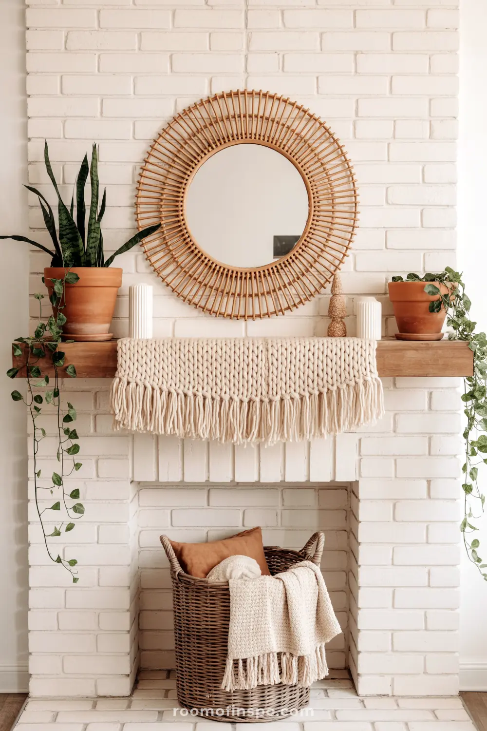A white brick fireplace features a rustic wooden mantel adorned with a woven macrame garland, potted plants, and a round rattan mirror.