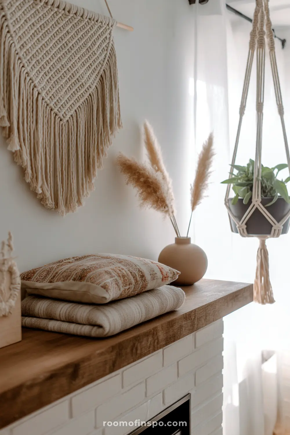 A cozy, rustic wooden mantelpiece is styled with a vase of pampas grass, stacked throw pillows, and a textured macrame wall hanging.