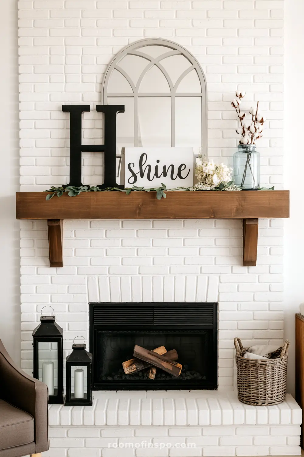 A charming white brick fireplace with a rustic wooden mantelpiece showcases an arched mirror, an "H" initial, a "Shine" sign, and cotton sprigs.