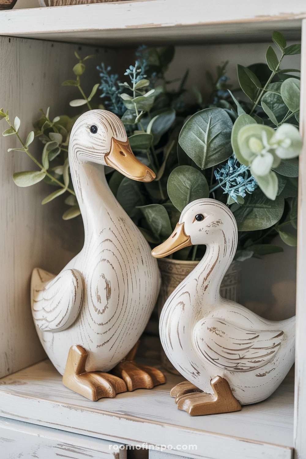 Two distressed white wooden duck figurines on a shelf with eucalyptus leaves and soft blue floral accents.