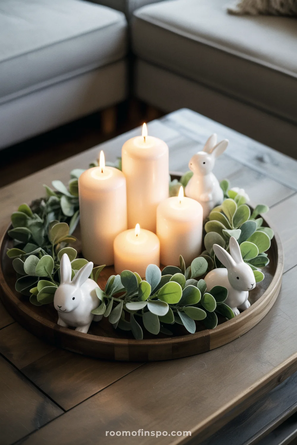 Round wooden tray centerpiece with white pillar candles, faux greenery wreath, and small ceramic bunny figurines.