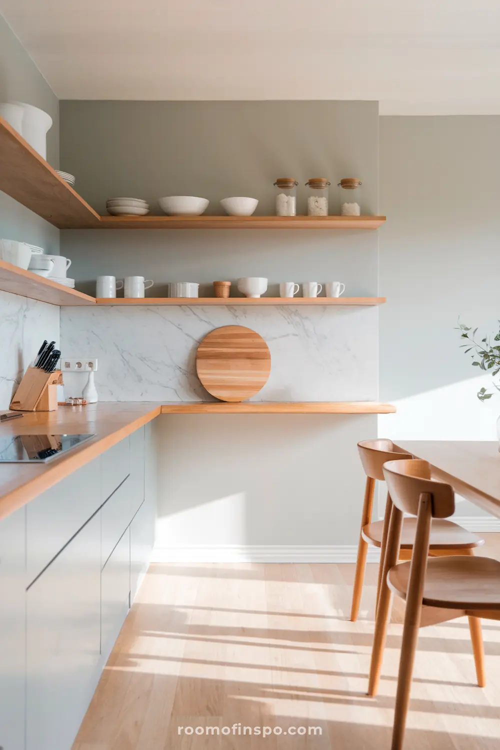 A modern Scandinavian kitchen with light wooden shelves holding white dishes and jars, a marble backsplash, a wooden cutting board, a knife block, and a dining table with wooden chairs.