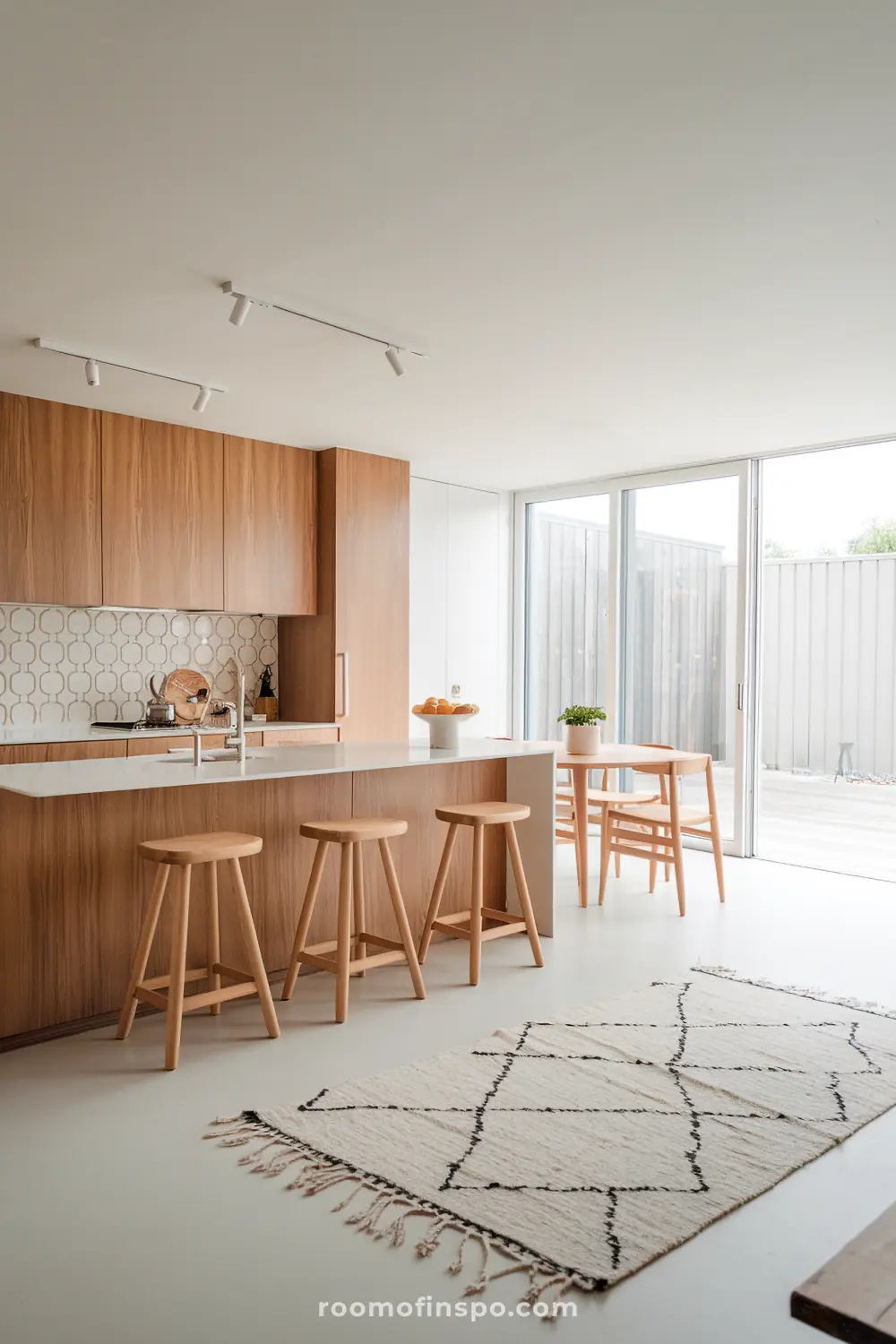 A modern Scandinavian kitchen and dining area with minimalist design, featuring wooden cabinets, a white countertop island with a built-in sink and faucet, wooden bar stools, a small dining table with two chairs and a potted plant, large sliding glass doors, a light-colored rug, and track lighting.