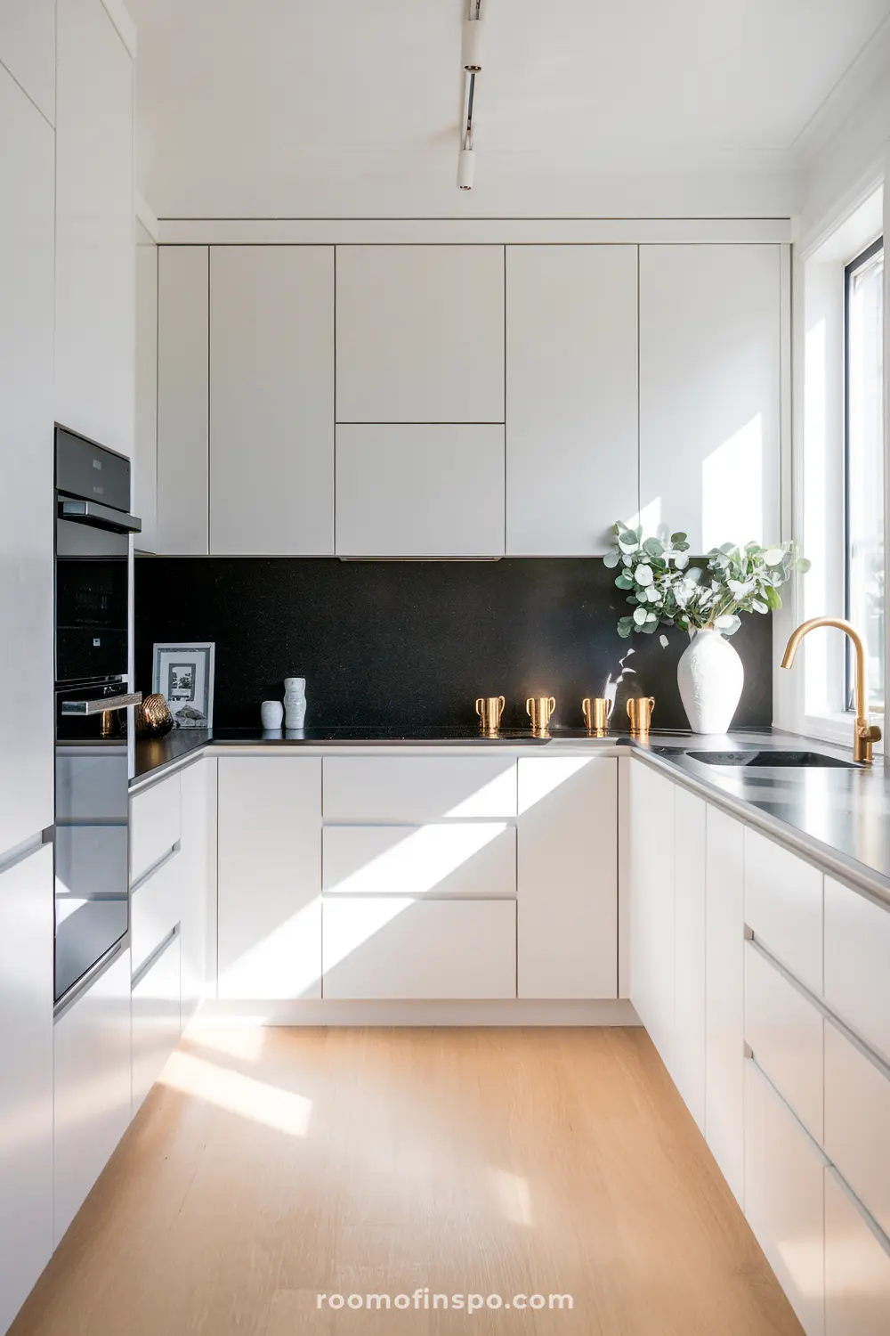 A modern Scandinavian kitchen with white cabinetry, black countertops, various decorative items, a sink with a gold faucet, and natural light from a window on the right side, with the text "roomofinspo.com" at the bottom.