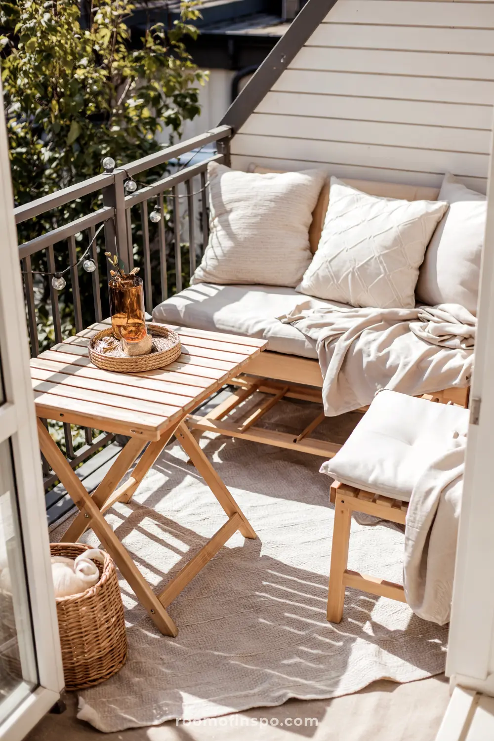 A small balcony featuring a cushioned bench with pillows and a blanket, a wooden table holding a tray with decor, string lights along the railing, and a view of trees and sunlight.
