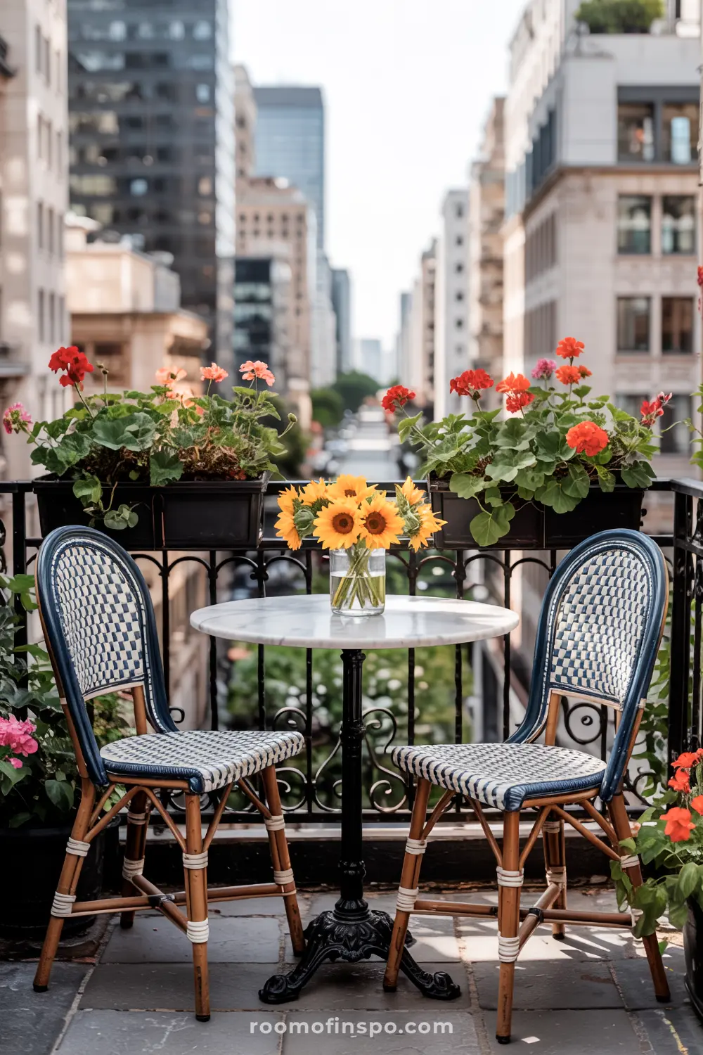 A small balcony with a round marble table featuring a bouquet of sunflowers, two wicker chairs with patterned cushions, flower boxes on the railing with vibrant red geraniums, and a cityscape in the background.