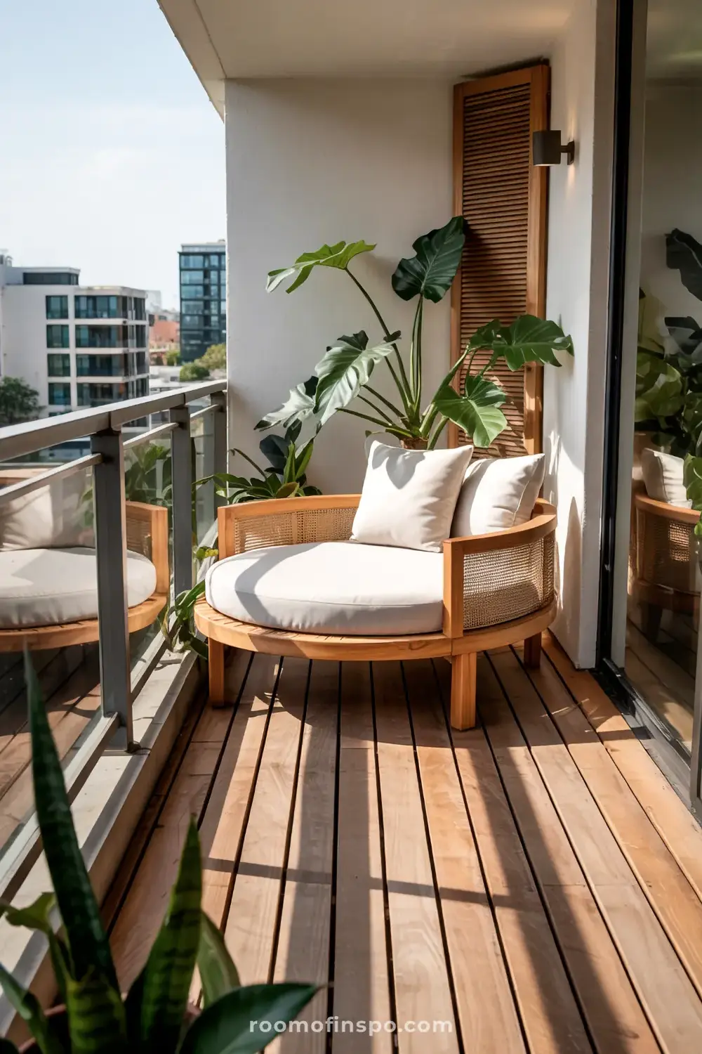 A compact balcony with wooden flooring, featuring a round rattan chair with cushions, large potted plants, and a glass railing, overlooking a cityscape.
