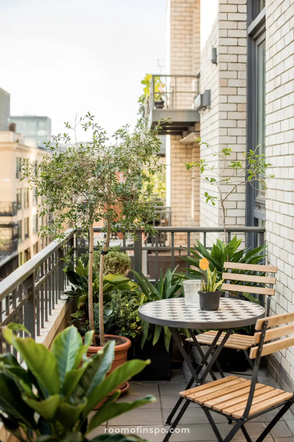 A compact balcony with a round checkered table, two wooden chairs, metal railing, and various potted plants, featuring a yellow-flowered plant and urban surroundings.