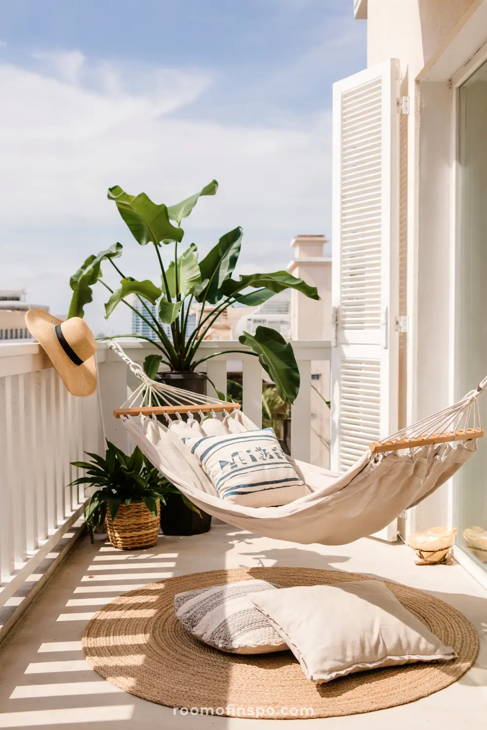 A small balcony with a hammock decorated with pillows, surrounded by potted plants and a woven rug on the floor, featuring a railing adorned with a straw hat and white shutters.