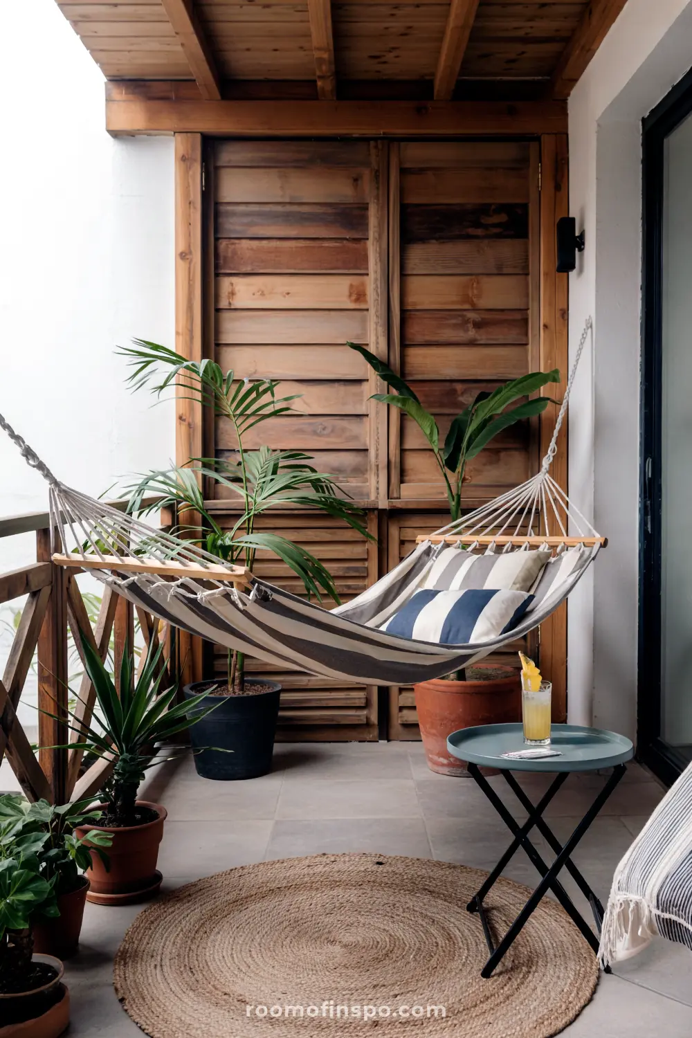 A compact balcony with a striped hammock, surrounded by potted plants, a glass-top table with a drink, and wooden sliding doors leading to the interior.