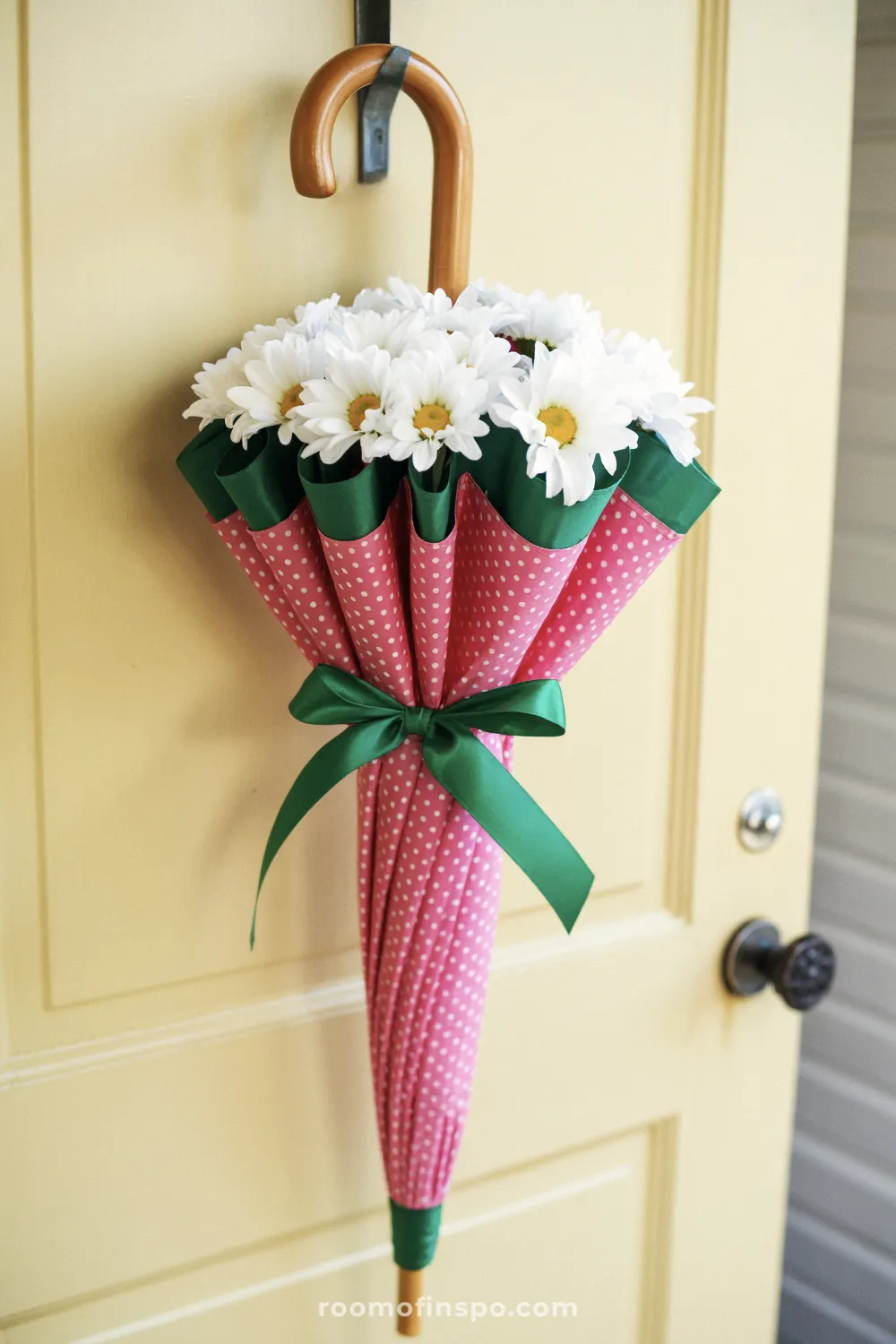 A pink umbrella with white polka dots is used as a vessel for white flowers, hanging on a cream colored door.