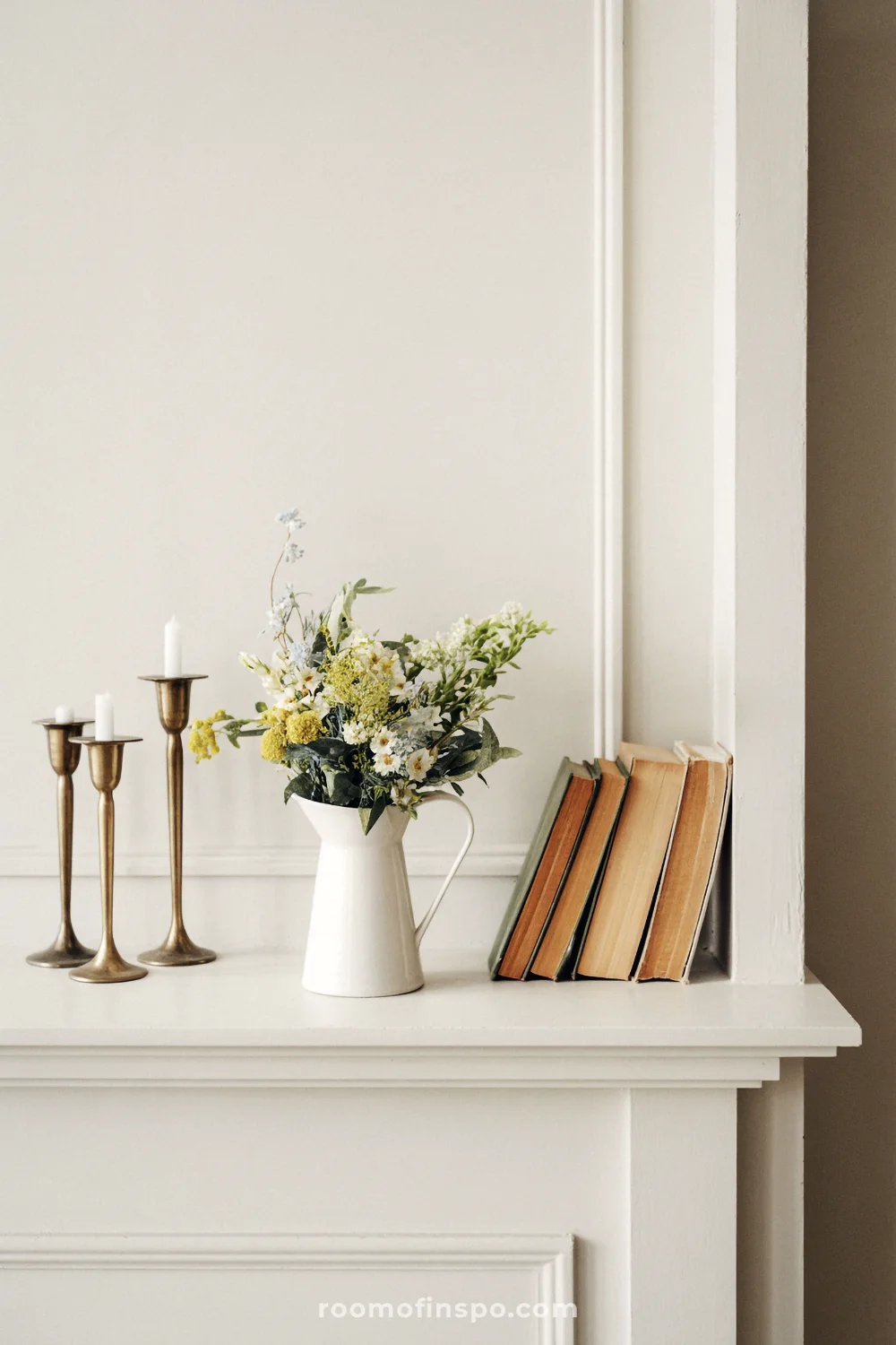 Minimal white mantel with small flowers, brass candlesticks, and neutral books against paneled wall.