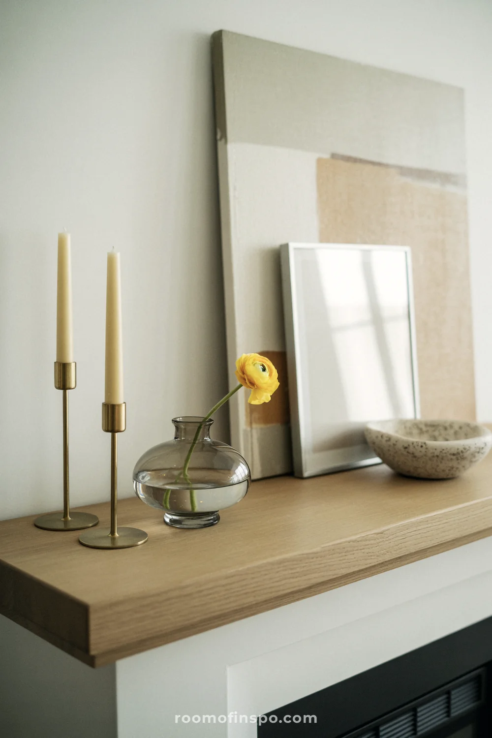 Floating oak mantel with brass tapers, a single yellow flower in a bud vase, and layered neutral art frames.