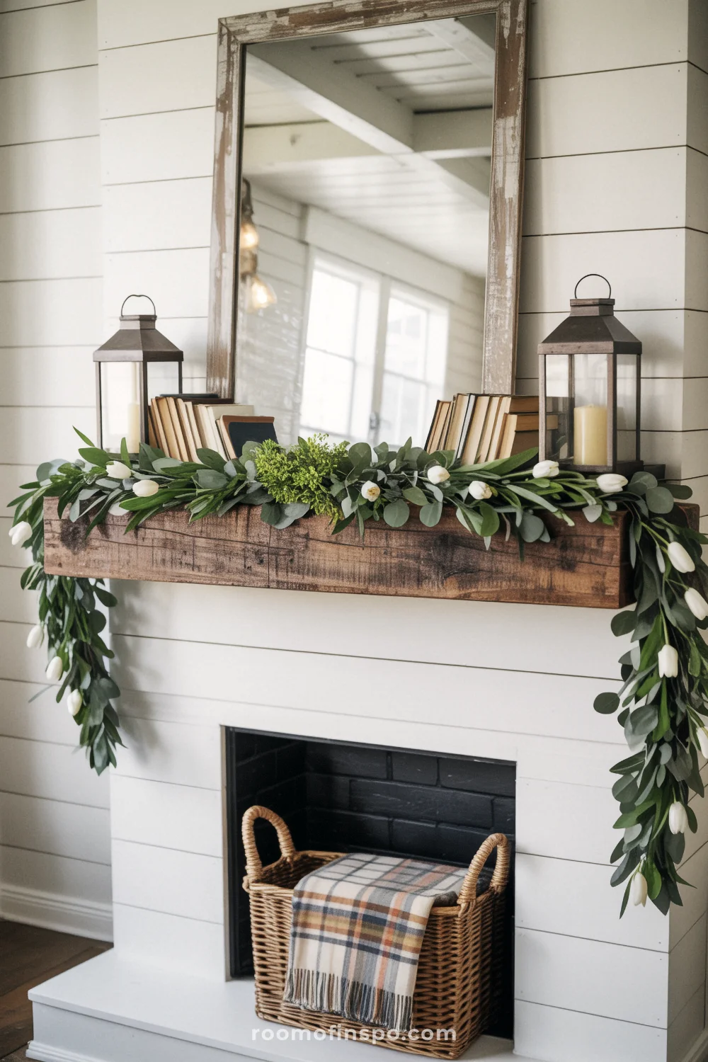 Reclaimed-wood mantel with eucalyptus garland and black lanterns beneath a distressed mirror on a white shiplap wall.