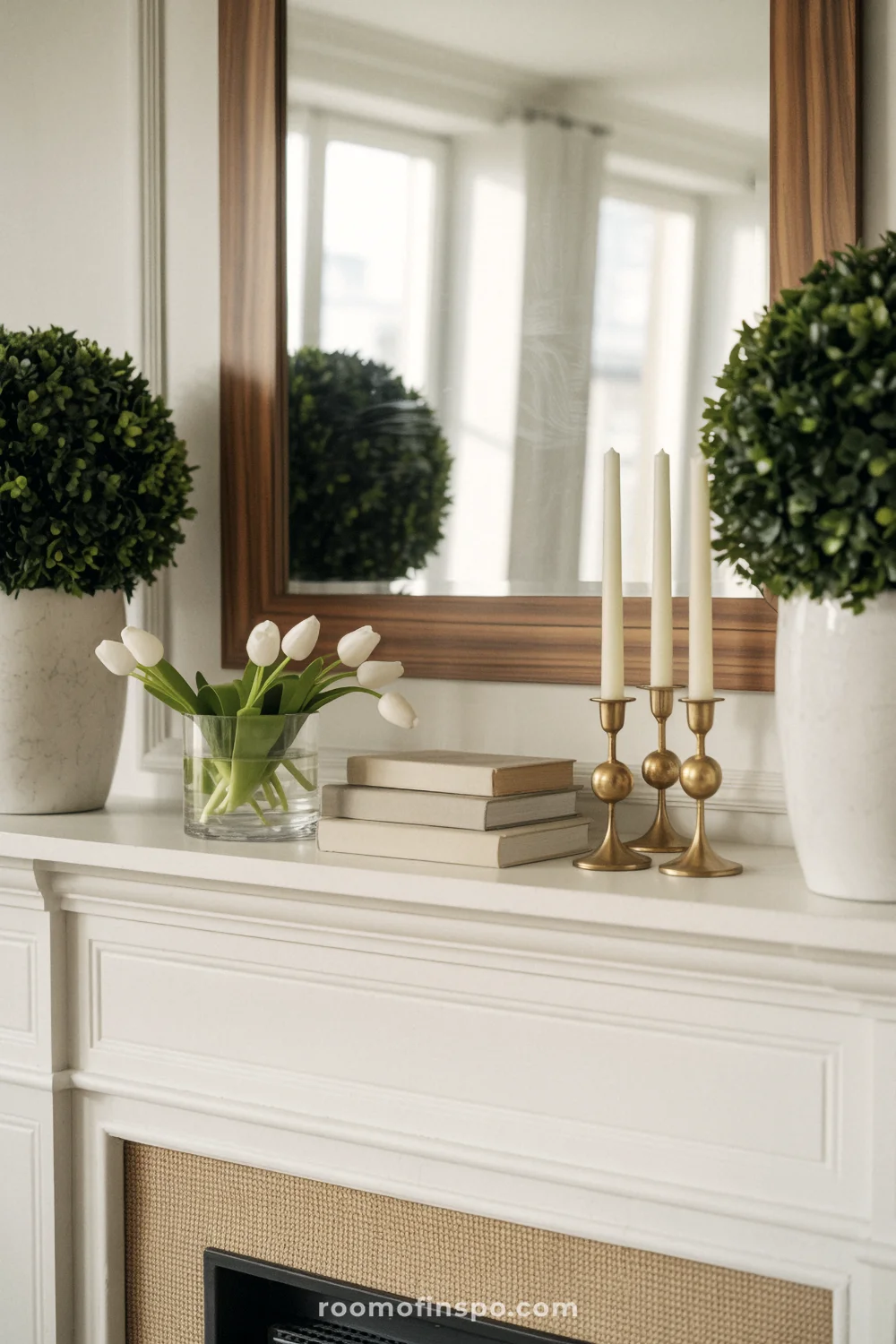 White mantel with two matching boxwood topiaries, a small tulip vase, brass candlesticks, and neutral books under a wood mirror.