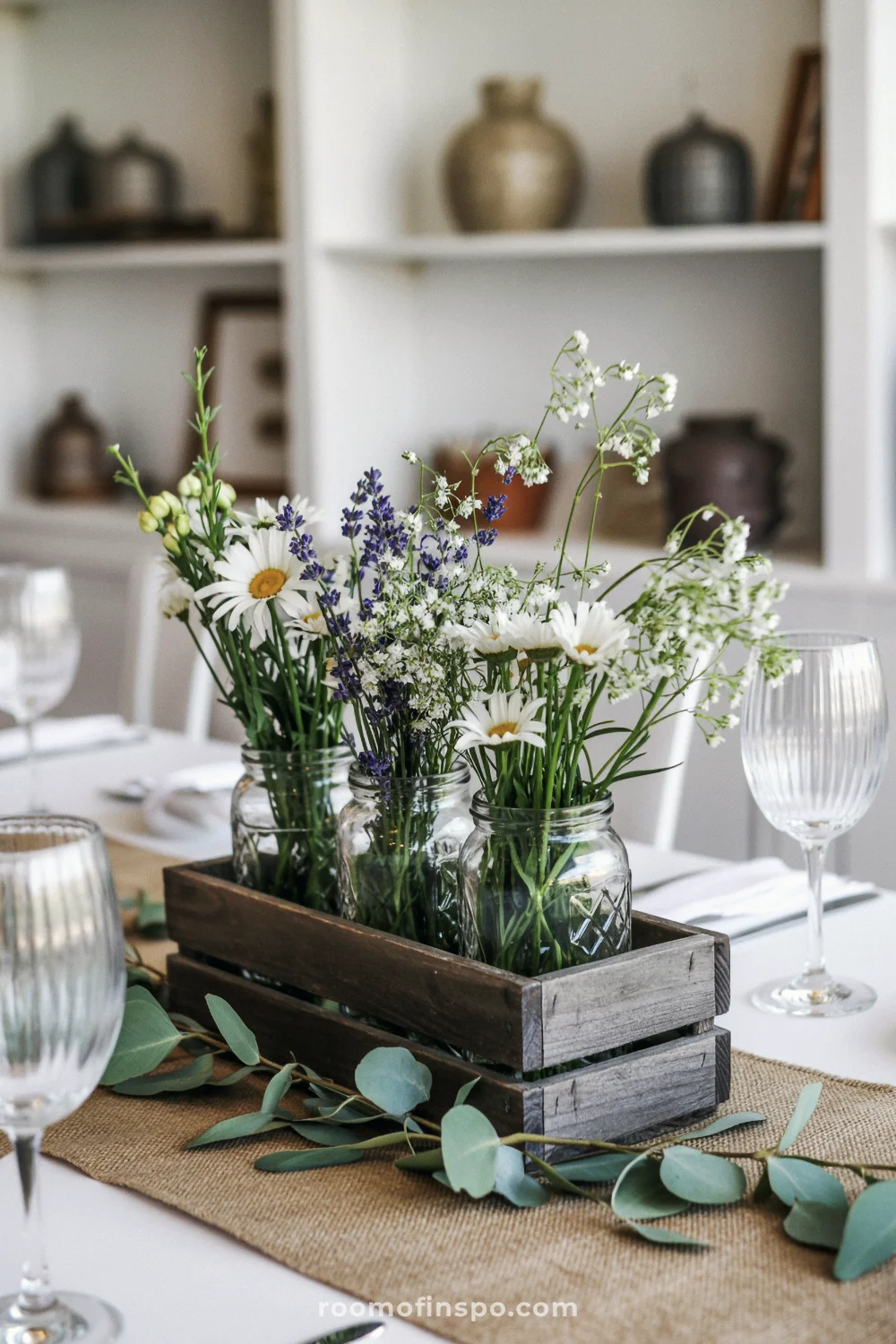 Rustic spring centerpiece with wildflowers in mason jars inside a wooden crate on burlap runner, styled with soft farmhouse decor.