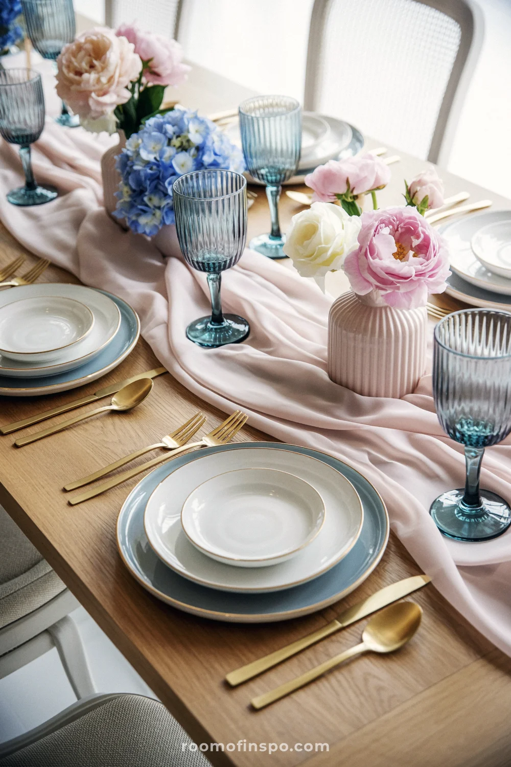 Pastel table with blush runner, blue chargers, gold flatware, small floral vases, and blue goblets.