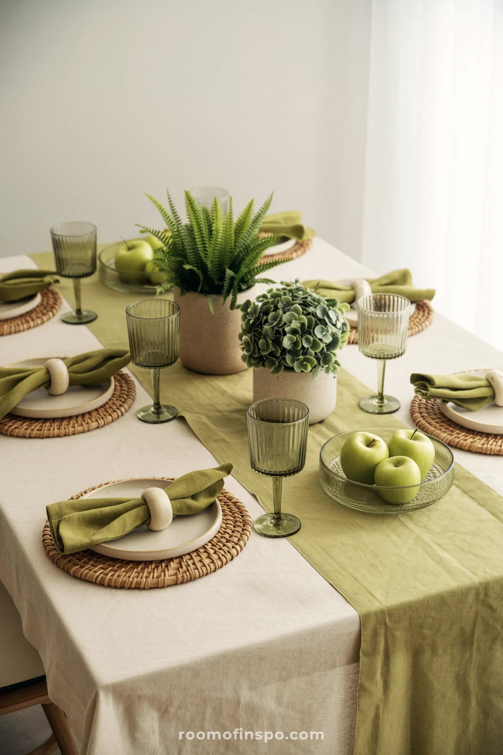 Green goblets and potted plants on a neutral linen table with rattan chargers and olive napkins.