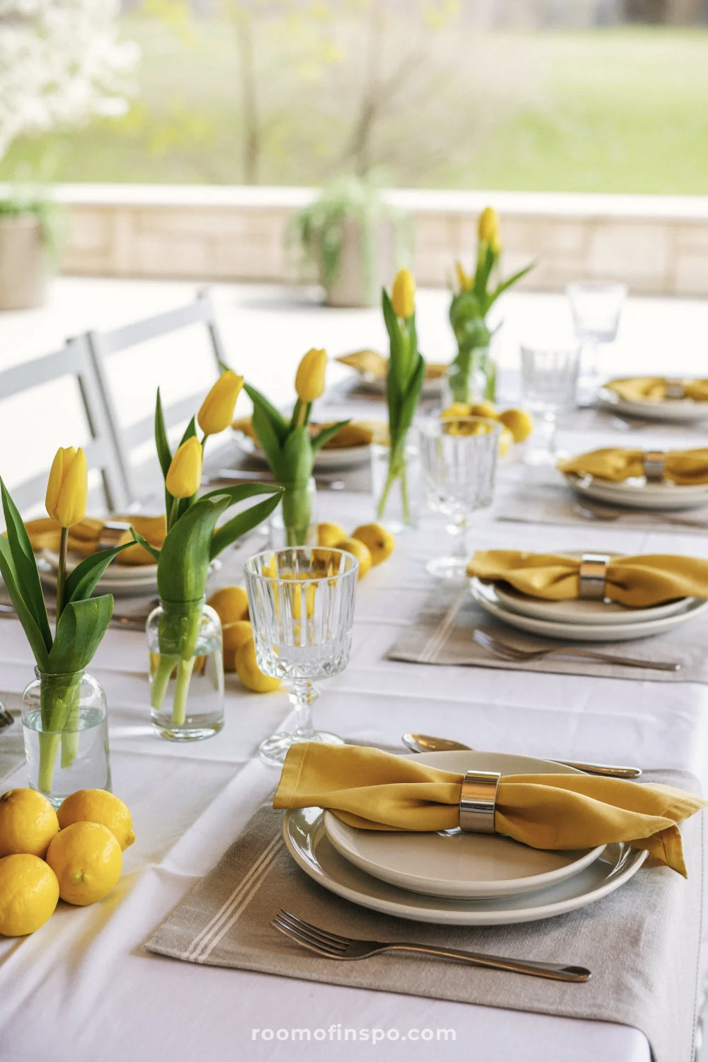 Patio table with white cloth, yellow tulips in bud vases, whole lemons, mustard napkins, and clear glassware.