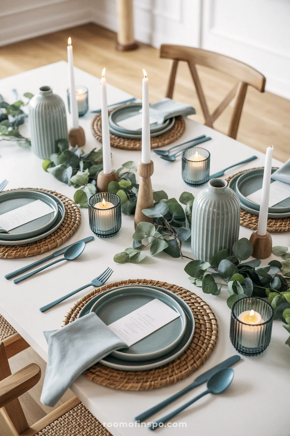 Coastal spring table setting with sage plates, eucalyptus, blue vases, and candles on white tablecloth in a bright, airy room.