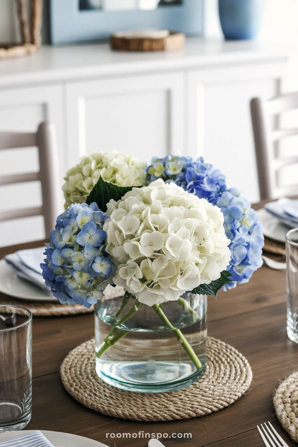 Spring coastal centerpiece with white and blue hydrangeas in a glass vase on dark wood table with bright natural lighting.