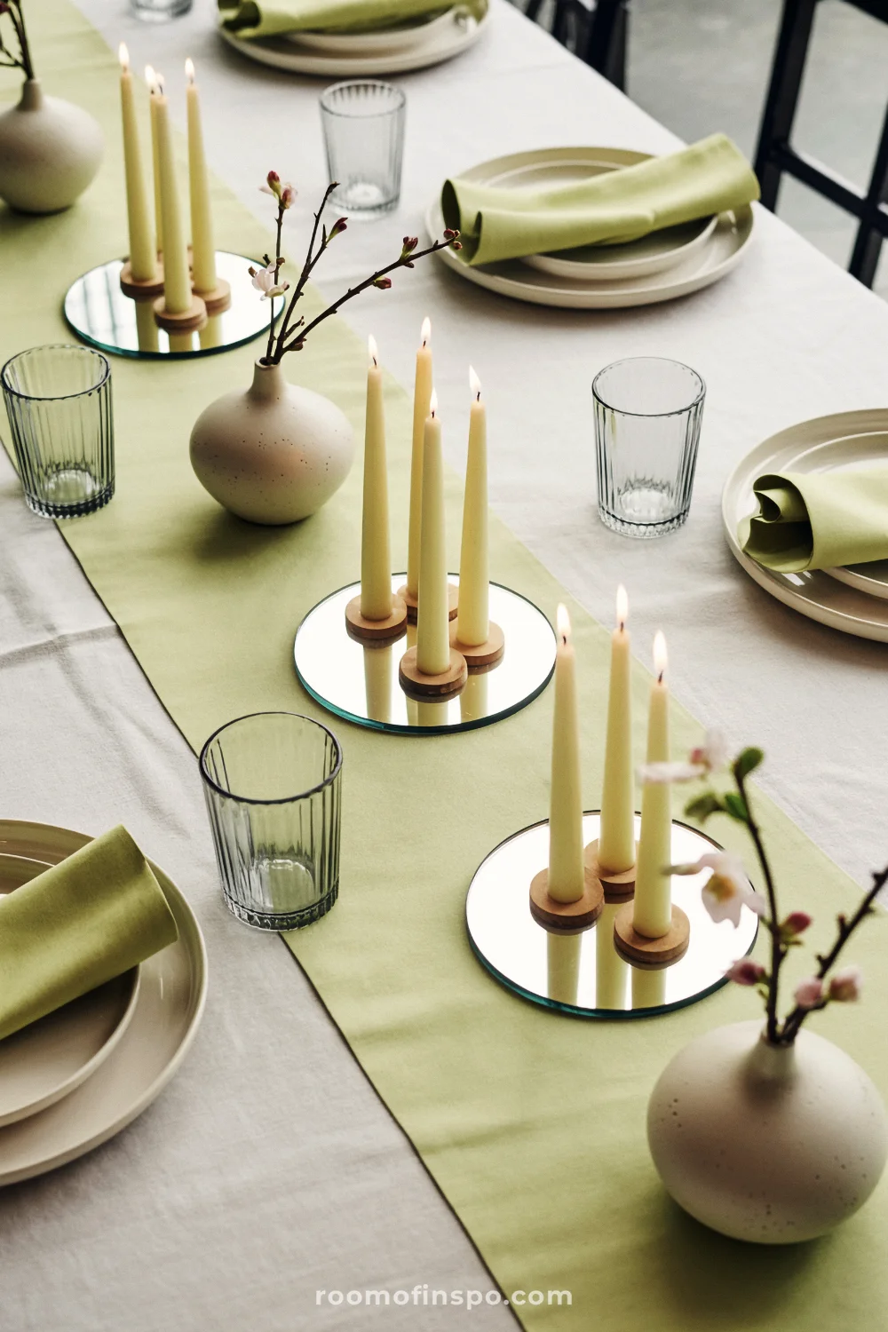 Clusters of slim candles on mirrored coasters with small bud vases on an ivory-and-celadon table setting.