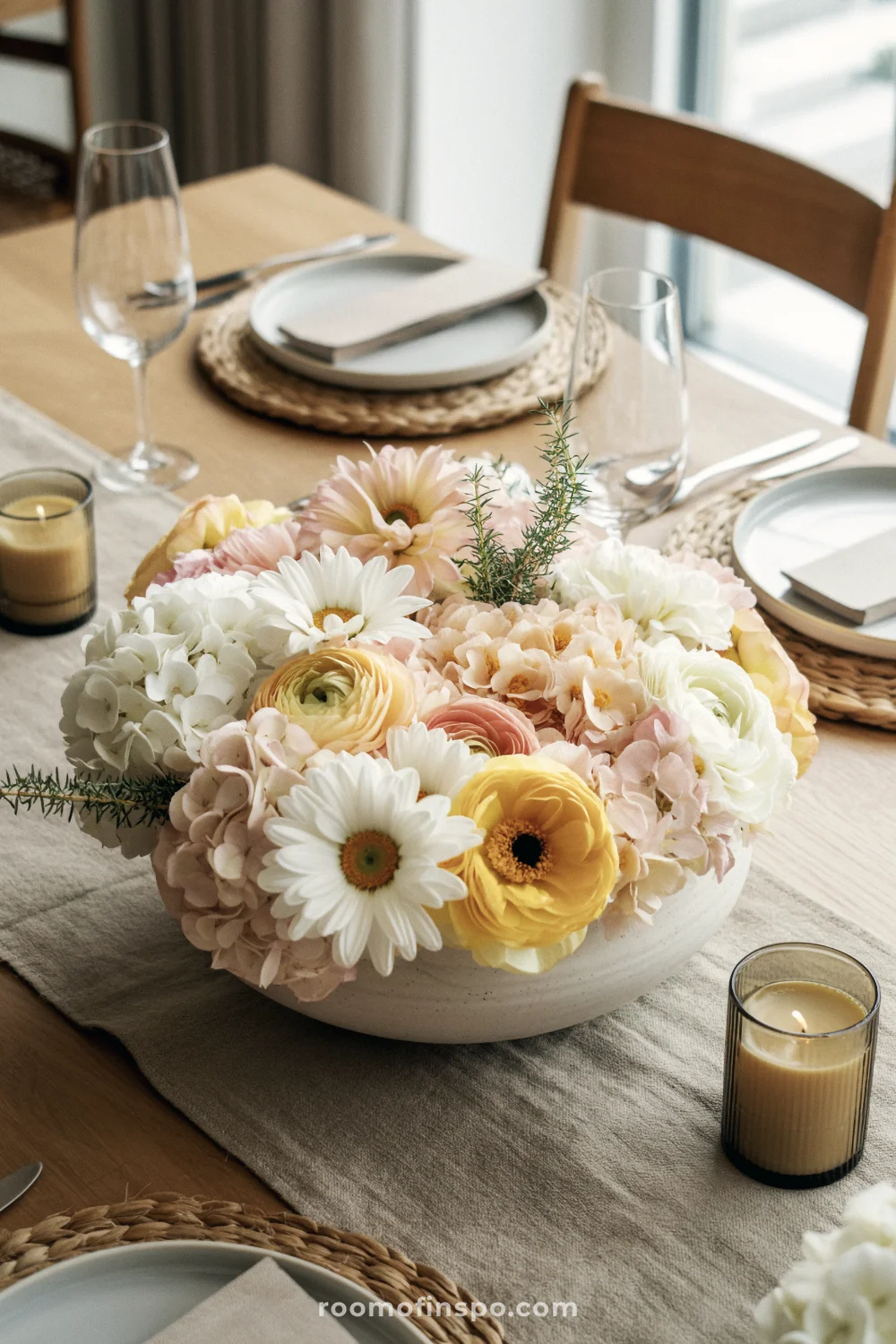 Low flower centerpiece in a wide bowl on a light wood table with white plates and clear glassware.