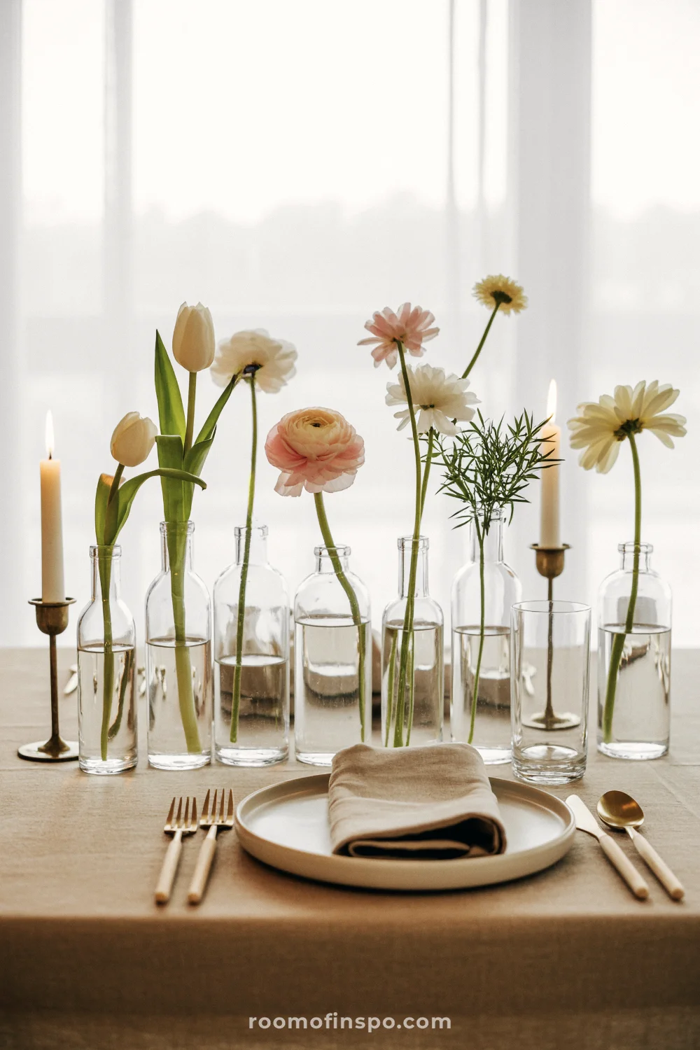 Line of small clear bottles with mixed spring stems on a neutral linen table with white dishes.
