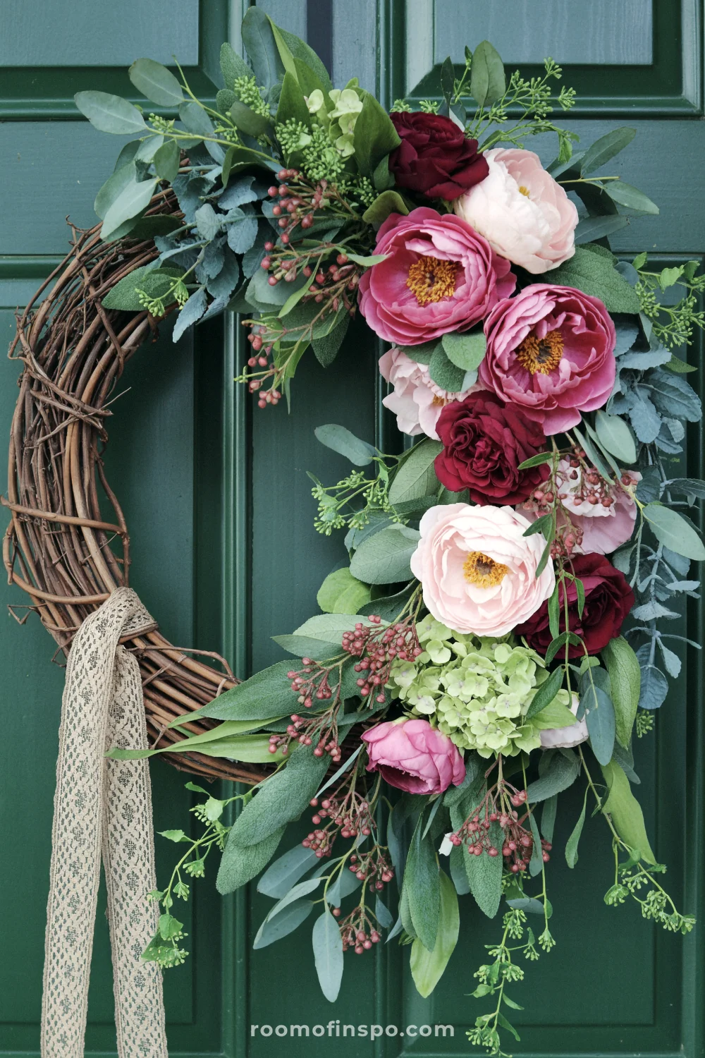 Cascading grapevine floral wreath with pink peonies, burgundy roses, greenery, and lace ribbon on dark green door.