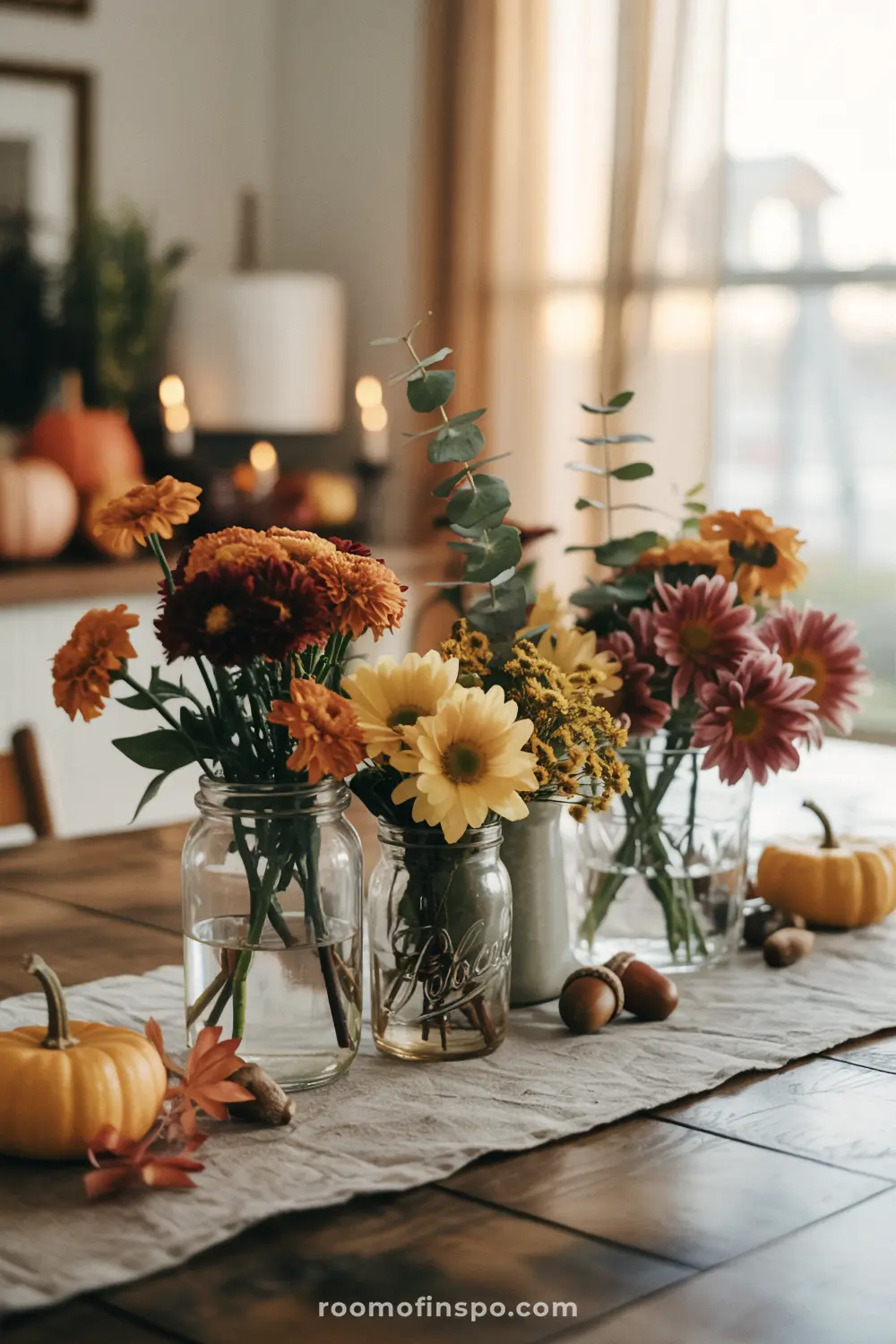 A cozy Fall centerpiece featuring a table runner with pumpkins, acorns, and assorted flowers in glass jars.