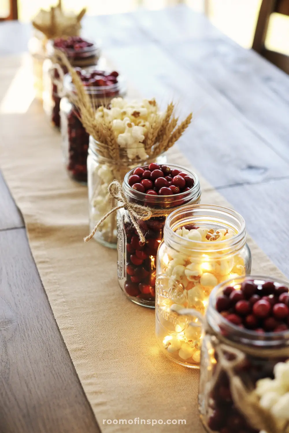 A unique Fall centerpiece with a row of mason jars filled with cranberries, popcorn, and fairy lights on a burlap runner.