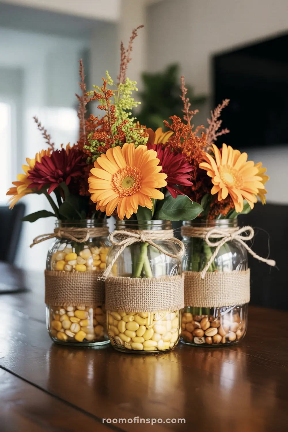 A lovely Fall centerpiece of three mason jars filled with assorted corn and beans, holding colorful autumn flowers.