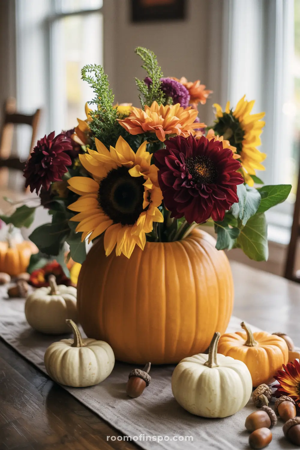 A festive Fall centerpiece with a pumpkin vase holding a bouquet of sunflowers and dahlias, surrounded by gourds and acorns.