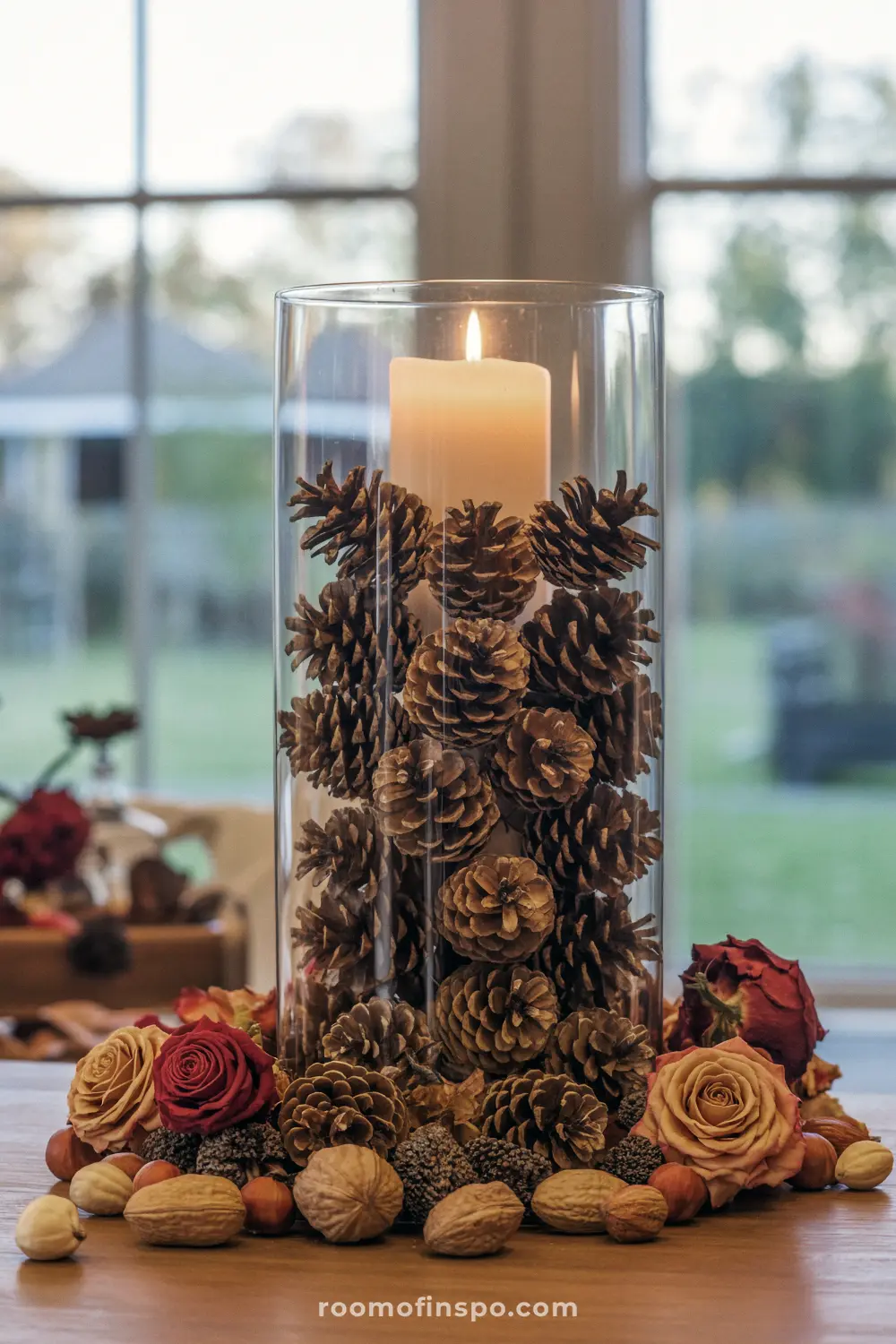 A rustic Fall centerpiece with a lit candle inside a glass hurricane vase, filled with pine cones and surrounded by nuts and dried flowers.