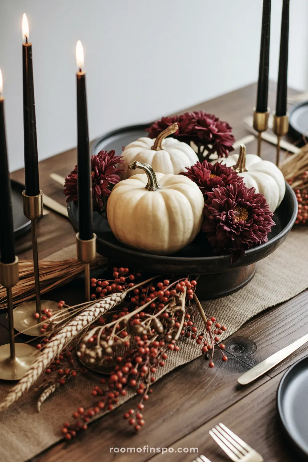 A centerpiece for a thanksgiving table with white pumpkins, deep red flowers, berries, and tall black candles.