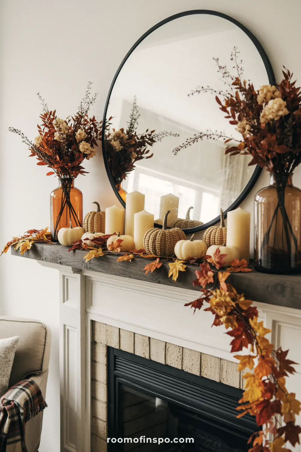 A fireplace mantel decorated for thanksgiving with a round mirror, fall leaves, gourds, candles, and tall vases.