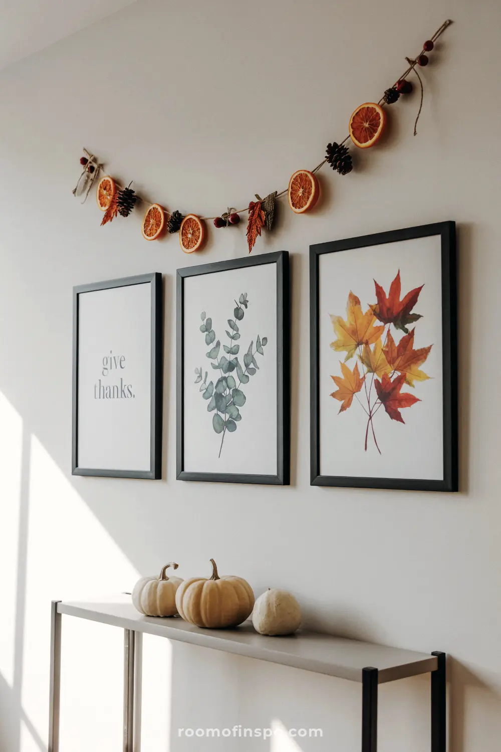 A modern thanksgiving wall display with a "give thanks" sign, a maple leaf print, and an orange slice garland.
