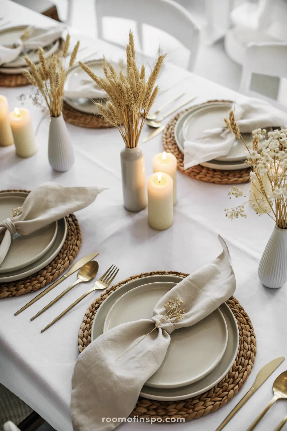 A white thanksgiving table with woven placemats, gold utensils, and vases of dried wheat.