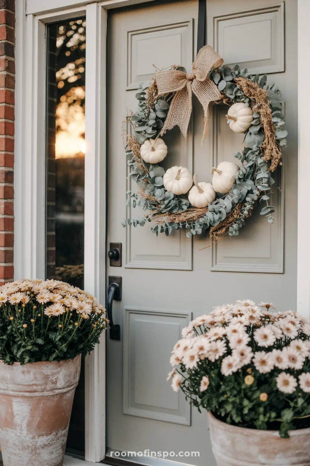A light-colored door with a thanksgiving wreath adorned with white pumpkins and a burlap bow.