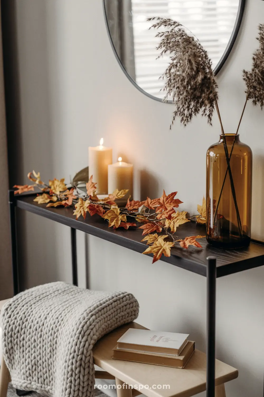 A console table decorated for thanksgiving with lit candles, a maple leaf garland, and dried grasses in a brown vase.