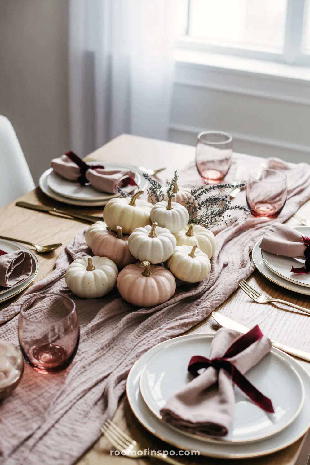 A Thanksgiving table centerpiece with a light pink runner, blush-colored glasses, and a pile of small white and pink pumpkins.