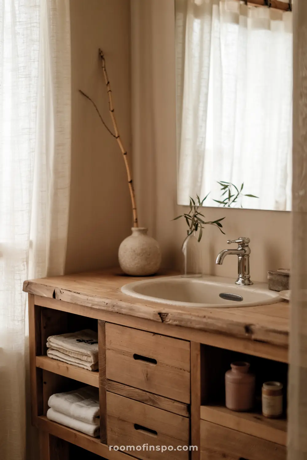 Wabi Sabi bathroom featuring a rustic wooden vanity with a white ceramic sink, a silver faucet, a round vase with a bamboo stick, neatly folded towels on open shelves, and natural light from a window with sheer white curtains.
