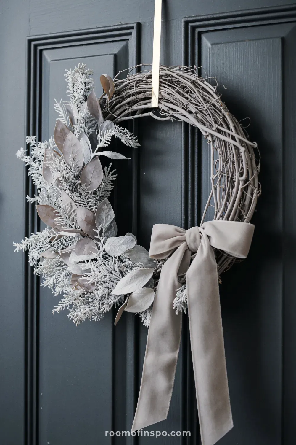 Rustic winter decor wreath featuring frosted foliage and a beige velvet bow hanging on a dark door.