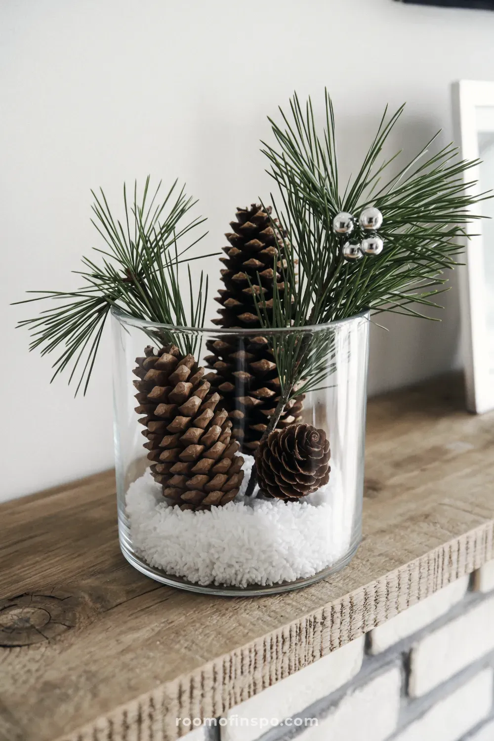 A glass cylinder displaying simple winter decor featuring pinecones, faux snow, pine needles, and silver berry accents.