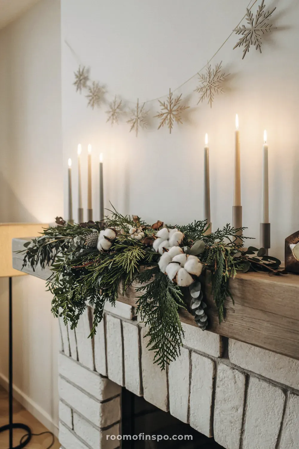 A warm mantlepiece featuring natural winter decor with greenery, cotton bolls, tall tapered candles, and a snowflake garland.