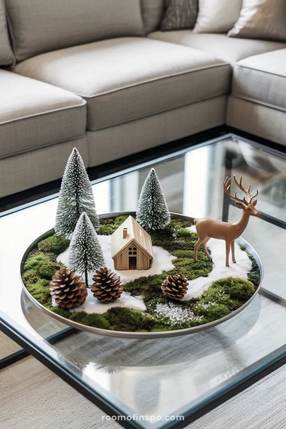 A glass coffee table features creative winter decor in a tray with tiny frosted trees, a miniature deer, and pinecones on a moss base.