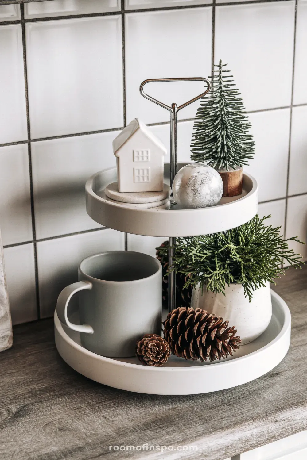 A two-tiered tray showcases neutral winter decor with pinecones, a mini house, a ceramic mug, and small potted greenery.