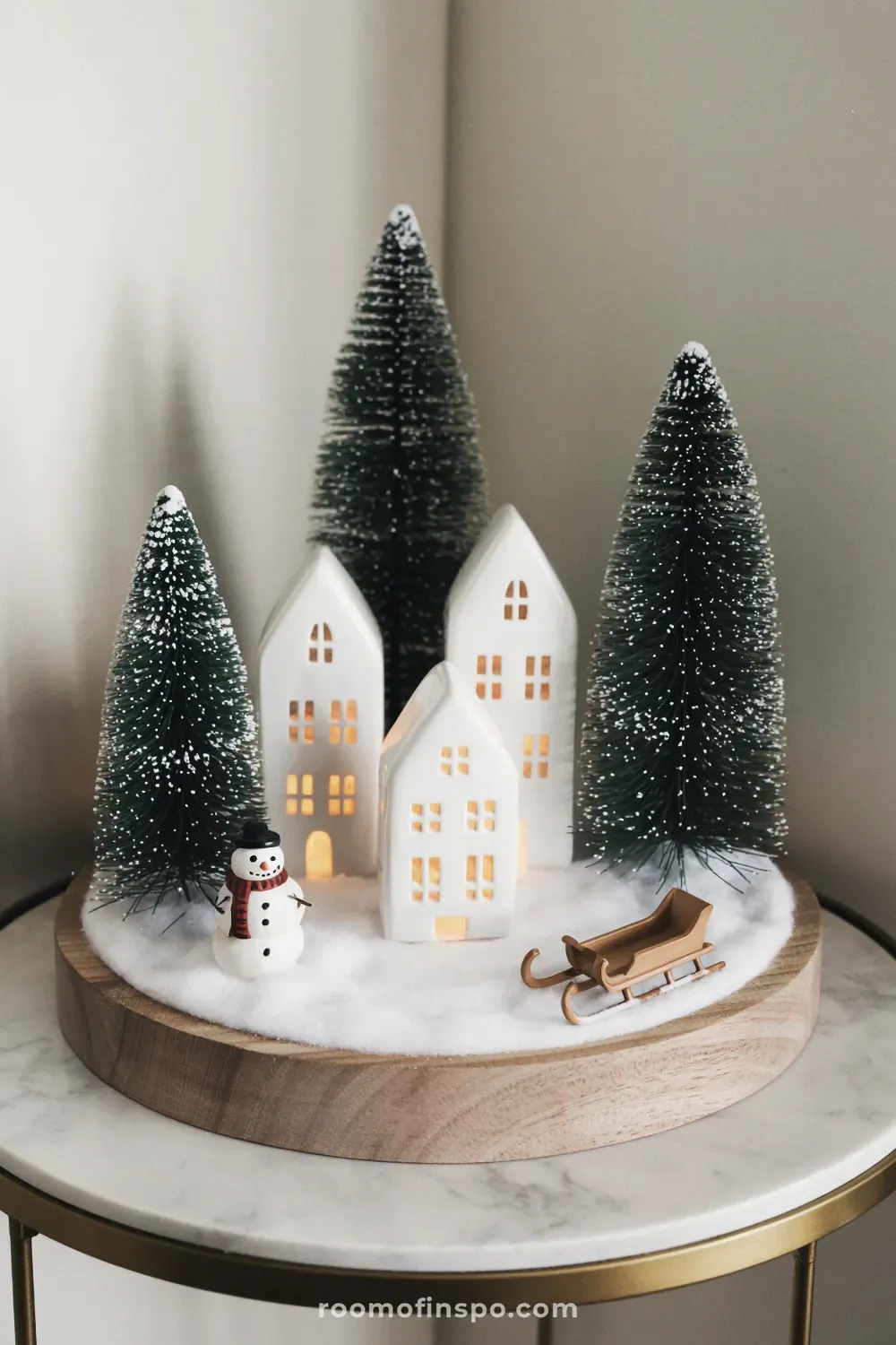 A marble table features illuminated winter decor with miniature white ceramic houses, bottle brush trees, and a small snowman.