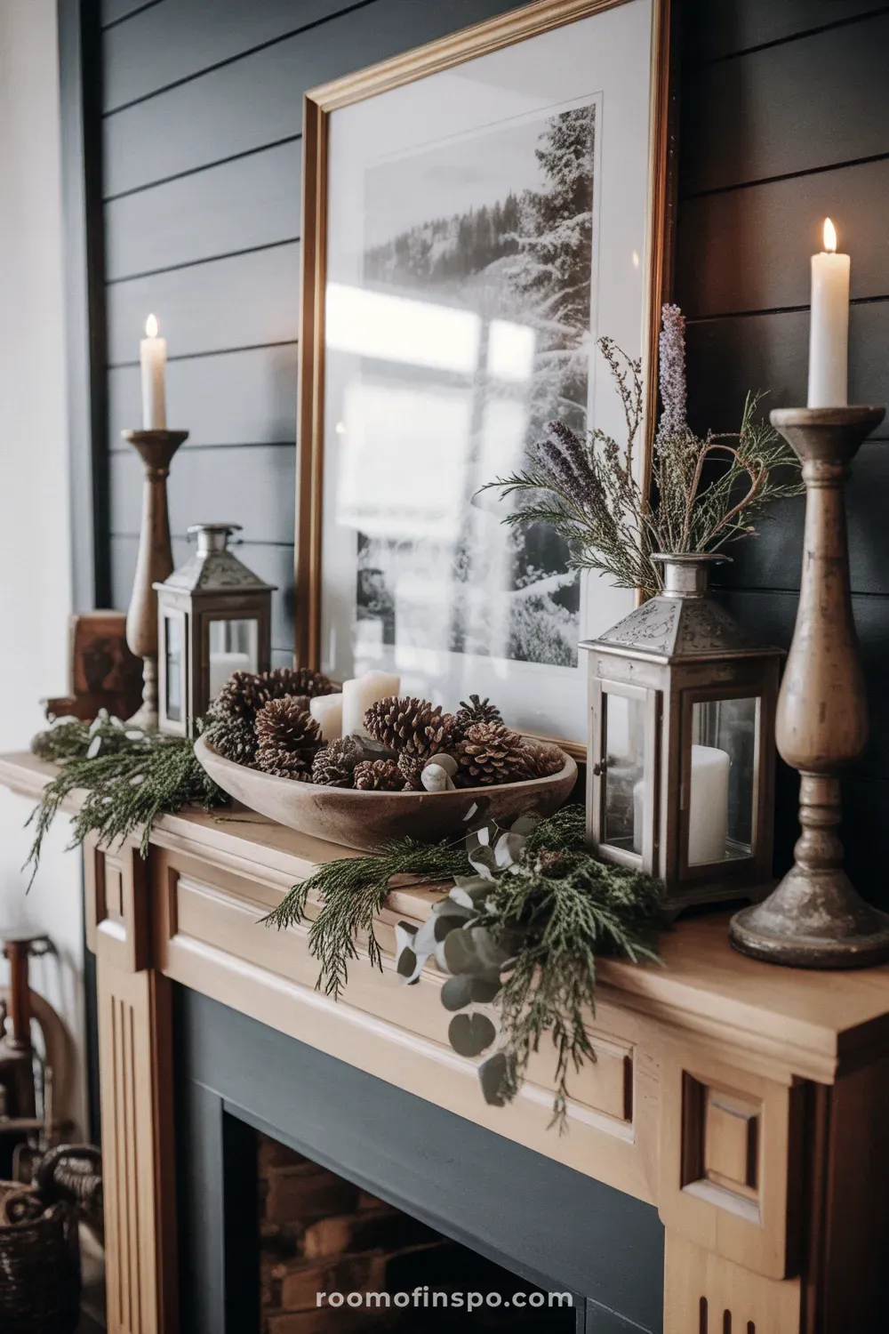 A dark, moody winter mantel display featuring pine branches, candles, and deep-toned accents around a picture.